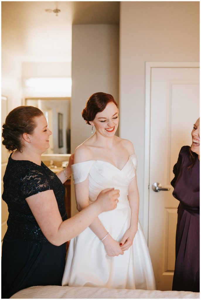 sister and mom help bride get ready on wedding day, laughing together