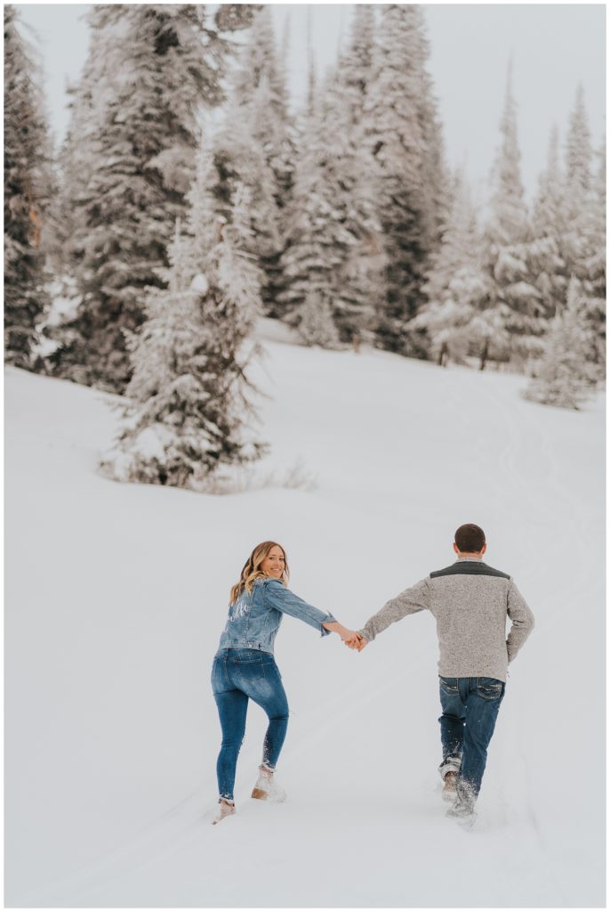 winter engagement session in twin falls idaho, south hills near magic mountain, bride and groom walking in snowy forest, bride looking back over her shoulder